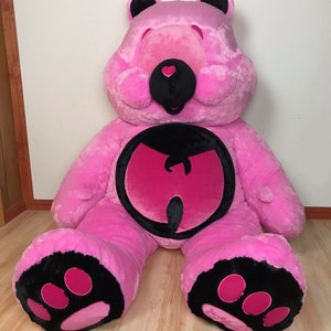May include: A large pink plush teddy bear with black ears and a black nose. The bear has a pink heart on its nose and a black Wu-Tang Clan logo on its chest. The bear's paws are black with pink paw pads.