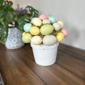 May include: A white ceramic pot filled with speckled pastel Easter eggs and green moss. The pot is sitting on a wooden table.