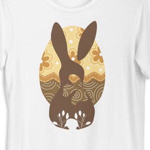May include: White t-shirt with a brown bunny rabbit silhouette inside a patterned Easter egg. The egg is decorated with brown, yellow, and white swirls and dots.