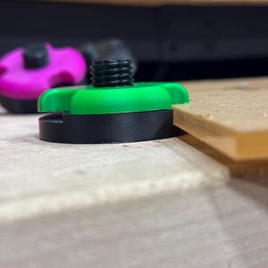 May include: A green and black 3D-printed component with a threaded post and a wing-shaped top. The component is resting on a wooden surface, with a blurred pink component in the background.