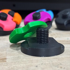 May include: Close-up of several colorful, 3D-printed objects. The objects have a black base with a threaded post and a colorful top. The tops are pink with orange, purple with blue, and green. The objects appear to be components or parts.