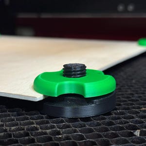 May include: Close-up of a laser cutter bed with a piece of light-colored wood supported by green and black leveling feet. The feet have a threaded post and a flower-shaped top. The bed has a honeycomb pattern.