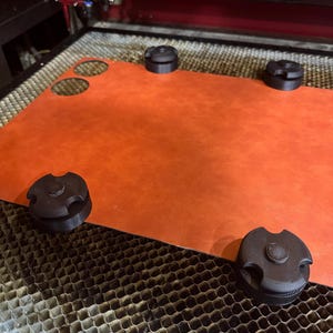 May include: An orange leather sheet with two circular cutouts is held in place by four black, circular clamps. The sheet is on a laser cutter bed. The laser cutter is in the background.