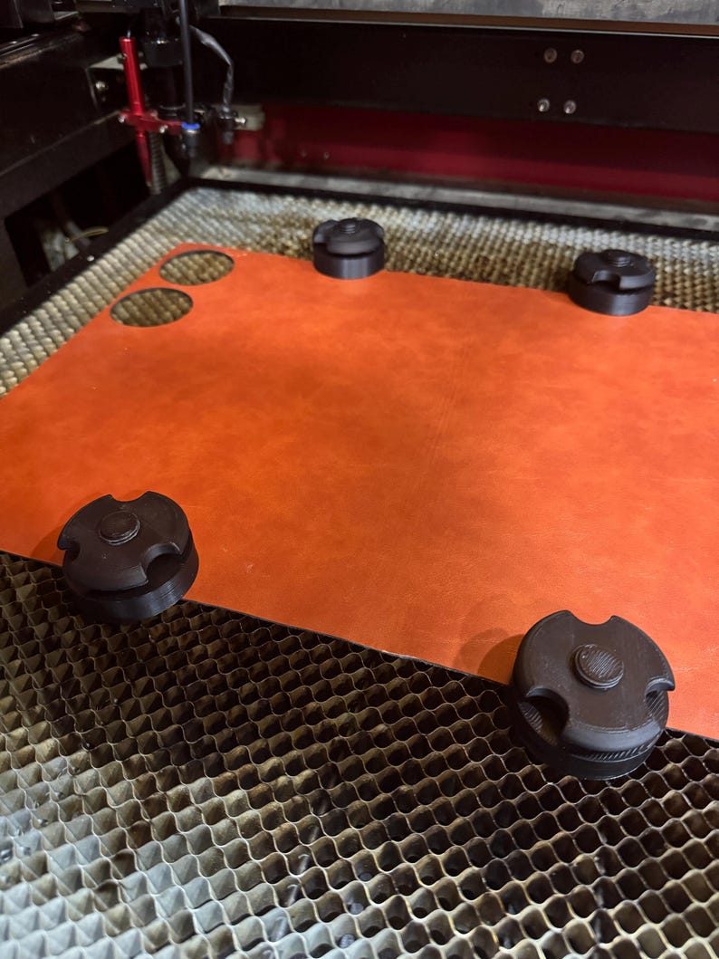 May include: An orange leather sheet with two circular cutouts is held in place by four black, circular clamps. The sheet is on a laser cutter bed. The laser cutter is in the background.