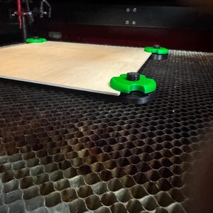May include: A light-colored wooden board is held in place by four green and black supports within a laser cutting machine. The machine has a black honeycomb-patterned base.