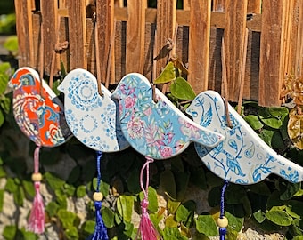 May include: Four ceramic bird ornaments hanging from brown leather cords. Each bird features a unique pattern: orange paisley, blue mandala, pink floral, and blue botanical. Each ornament includes a small wooden bead and a colorful tassel.