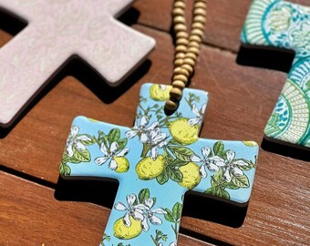 Puede incluir: Un adorno de cerámica en forma de cruz con un fondo azul claro y un patrón de limones amarillos, hojas verdes y flores blancas. El adorno está suspendido por un cordón de pequeñas cuentas de madera de color marrón claro.