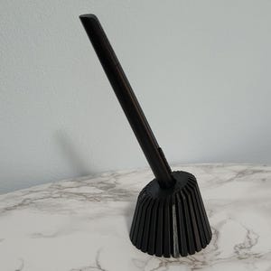 May include: A dark brown bottle stopper with a long, slender handle and a flared, ribbed base. The stopper is made of wood and is designed to fit into a bottle. The base has vertical grooves.
