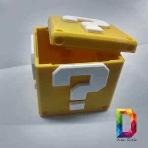 May include: A yellow 3D printed box with a white question mark on the front. The box is open and the lid is off. The box is a replica of a question block from the Super Mario Bros. video game.