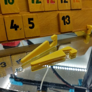 May include: Yellow plastic game tile holders with a wooden game board in the background. The wooden tiles have black numbers printed on them. The numbers 3, 4, 5, and 13 are visible. The holders are reflected in a mirrored surface.