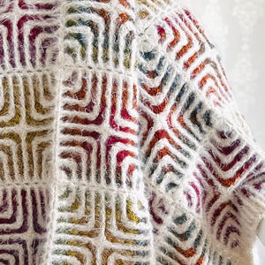 May include: A cream-colored, textured knit garment with a geometric pattern in shades of red, orange, yellow, and teal. The design features repeating square and angled shapes, creating a visually interesting texture. The garment appears soft and cozy.