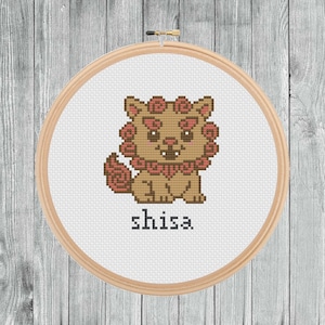 May include: A cross-stitch embroidery of a Shisa, a mythical creature, in a wooden hoop. The Shisa is brown with a pink mane and collar, and the word "shisa" is stitched below. The background is white.