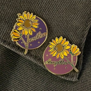 Puede incluir: Dos pines de esmalte con fondo morado y contornos dorados. Cada pin tiene un girasol amarillo con hojas verdes y una pequeña flor amarilla. El pin superior dice "madre" y el pin inferior dice "hija".