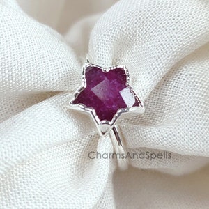 Peut inclure: Une bague en argent avec une pierre précieuse en forme d'étoile en rubis. La bague est posée sur un fond en tissu blanc. Le texte 'CharmsAndSpells' est imprimé sous la bague.