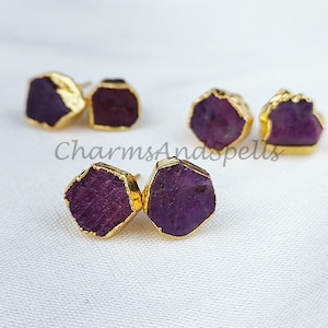 Puede incluir: Un par de pendientes chapados en oro con detalles de piedras preciosas púrpuras. Las piedras preciosas tienen una forma irregular y un aspecto áspero y natural.