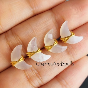 May include: Three white crescent moon shaped gemstone charms wrapped in gold wire. The charms are displayed on a hand.