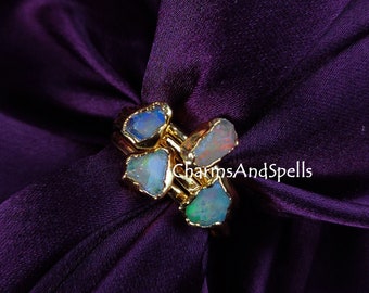 May include: A gold-colored ring featuring multiple raw opal stones in shades of blue, green, and white. The ring is set against a deep purple satin fabric. The text "CharmsAndSpells" is visible.