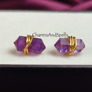 Może przedstawiać: Para kolczyków z ametystowymi kryształami owiniętymi złotym drutem. Kryształy ametystowe są ciemnofioletowe i mają kształt długiego, cienkiego szpiczastu. Złoty drut jest owinięty wokół kryształów w prosty, elegancki wzór.