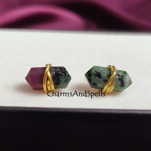May include: A pair of earrings featuring green and pink gemstones wrapped in gold wire. The earrings are displayed on a black surface.