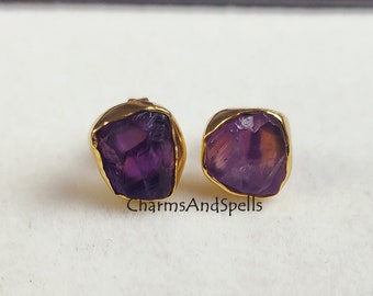 May include: A pair of gold stud earrings with purple amethyst gemstones. The earrings are set in a simple, modern style.