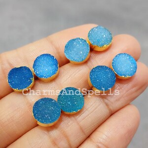 Può includere: Una collezione di gemme rotonde di druzy blu con bordi color oro. Le gemme variano nelle tonalità di blu, da chiaro a scuro. L'immagine è ben illuminata, mostrando la consistenza e la brillantezza delle pietre. Il testo "CharmsAndSpells" è visibile.