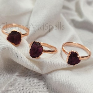 May include: Three rose gold rings with raw ruby stones. The rings are on a white fabric background. The text "CharmsAndSpells" is visible in the top left corner of the image.