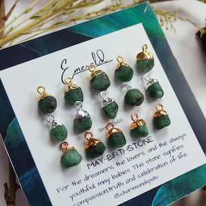 May include: Emerald green gemstone charms with gold accents. The charms are arranged on a white card with the text "Emerald" and "MAY-BIRTHSTONE" along with a description of the stone's meaning. The text reads: "For the dreamers, the lovers, and the always youthful may babies. This stone signifies compassion truth and celebration of life. @charmsandspells"