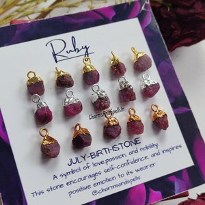 以下が含まれることがあります： 金と銀のアクセントが付いたルビーのチャーム12個のセット。チャームは、「Ruby」と「July-Birthstone」の文字が印刷された白いカードに並べてあります。テキストには、ルビーが愛、情熱、高貴さの象徴であることも記載されています。カードには「@charmsandspells」の文字も含まれています。