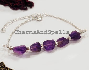 May include: A silver chain bracelet with a row of five purple amethyst stones. The bracelet has a lobster clasp closure. The text "CharmsAndSpells" is visible in the image.