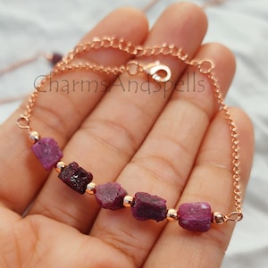 Peut inclure: Un bracelet en chaîne en or rose avec une rangée de petites pierres de rubis brutes. Le bracelet est sur une main, et le texte "CharmsAndSpells" est visible en arrière-plan.
