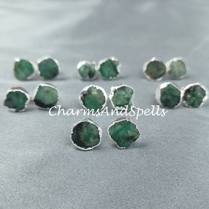 以下が含まれることがあります： エメラルドグリーンの宝石のイヤリング10個セット。各イヤリングは、シルバーのベゼルセッティングにセットされた、カットされていない生のエメラルドストーンが特徴です。イヤリングはグレーの布地に置かれています。