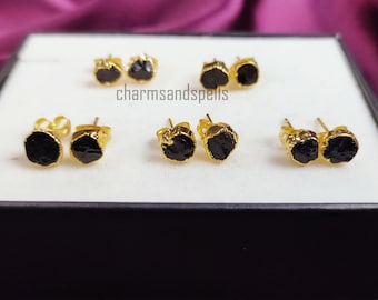 May include: Several pairs of black tourmaline stud earrings with gold-colored settings are displayed in a black box. The earrings have a raw, natural appearance, with irregular shapes and textures. The text "charmsandspells" is visible.