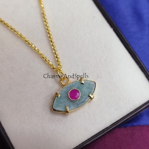 Puede incluir: Collar de cadena dorada con un colgante de ojo turco azul claro y dorado con una piedra preciosa rosa en el centro. CharmsAndSpells.