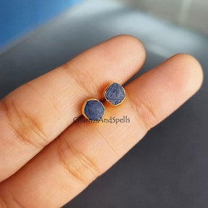 May include: A pair of gold stud earrings with small, square-shaped blue sapphire stones. The stones have a rough, natural texture.
