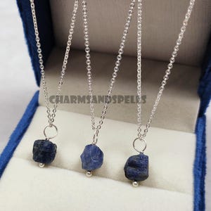 Peut inclure: Trois colliers en chaîne argentée, chacun avec un pendentif en saphir brut bleu foncé et une petite perle argentée. Les colliers sont présentés dans un écrin en velours bleu. Le texte "CHARMSANDSPELLES" est visible.