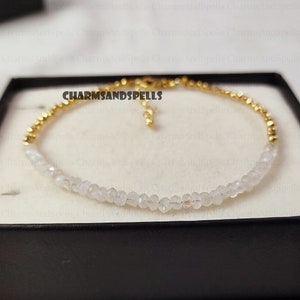 May include: A delicate bracelet made with white gemstone beads and gold accents. The bracelet is displayed in a black box with the text "CHARMSANDSPELLS" printed on the inside.