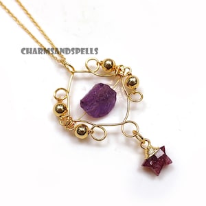Peut inclure: Collier doré avec un pendentif carré contenant une améthyste violette brute et un pendentif en forme d'étoile. Le collier présente des perles dorées et des détails en fil de fer. Le texte "CHARMSANDSPELLS" est visible en haut à gauche.