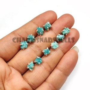 May include: A collection of raw, teal-colored Amazonite stud earrings. Each earring features a small, irregularly shaped gemstone held in a silver-colored metal setting. The earrings are displayed on a light background.