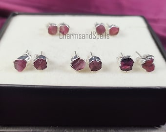 May include: Six pairs of silver earrings with pink, raw, crystal stones. The earrings are displayed in a black box with a white interior. The text "CharmsandSpells" is printed on the box.