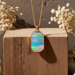 Puede incluir: Un collar de cadena dorada con un colgante rectangular. El colgante presenta una piedra de ópalo iridiscente con tonos de azul, verde y rosa. El collar se exhibe sobre un bloque de madera, con flores secas en el fondo.