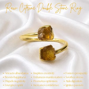 Puede incluir: Un anillo abierto de tono dorado con dos piedras de citrino en bruto de color ámbar en los extremos. El texto "Raw Citrine Double Stone Ring" aparece en la parte superior, con una lista de atributos que incluye "Attracts abundance," "Boosts happiness," e "Inspires creativity" impresos debajo.