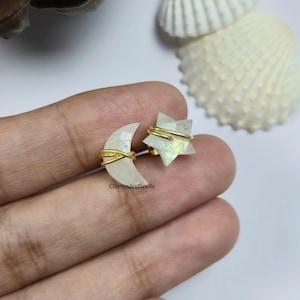 Pode incluir: Um par de brincos com tema celestial, com uma lua crescente e uma estrela. A lua e a estrela são brancas com fio dourado. O texto "CharmsAndSpells" é visível.