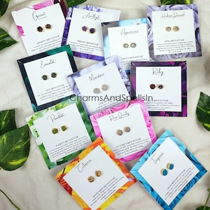 May include: A set of 12 pairs of gemstone earrings, each pair displayed on a card with the gemstone's name, birth month, and a brief description of its meaning. The cards are arranged in a grid pattern on a surface with green leaves.