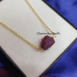 Può includere: Una collana con catena dorata e un ciondolo di rubino grezzo. Il rubino è di colore rosso intenso e ha una texture ruvida e naturale. La collana è esposta in una scatola nera con sfondo blu. Il testo "CharmsAndSpells" è visibile sulla scatola.