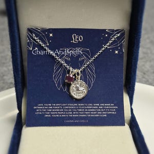 以下が含まれることがあります： レオの星座のチャームと小さな赤い宝石のペンダントが付いたシルバーネックレス。ネックレスは、青いベルベットの背景に、「LEO CharmsAndSpells」とレオの星座の説明が書かれたカードと共に飾られています。