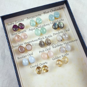 May include: A collection of 12 pairs of gemstone earrings displayed in a blue box. The earrings are arranged in two rows of six pairs. Each pair is labeled with the name of the gemstone. The gemstones include ruby zoisite, rose quartz, chocolate moonstone, boulder opal, crystal quartz, golden rutile, amazonite, aqua chalcedony, labradorite, black rutile dendrite opal, pink opal, and rainbow moonstone.