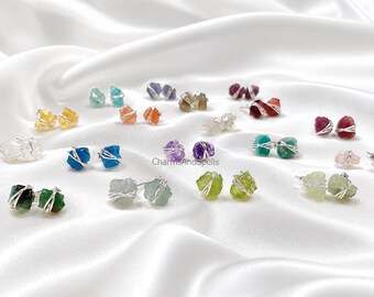 May include: A collection of raw gemstone stud earrings, each featuring a rough, unpolished crystal wrapped in thin silver-colored wire. The stones appear in various colors including blue, green, yellow, orange, purple, red, and clear.