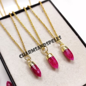Może przedstawiać: Trzy złote łańcuszki z zawieszkami w kształcie różowych kryształowych punktów. Naszyjniki są prezentowane w czarnym pudełku z widocznym tekstem "CHARMSANDSPELLS".