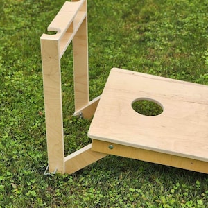 Può includere: Un set da gioco di cornhole in legno con finitura in legno naturale. Il set comprende due tavole con un foro circolare al centro e un supporto in legno con due staffe per tenere le tavole.