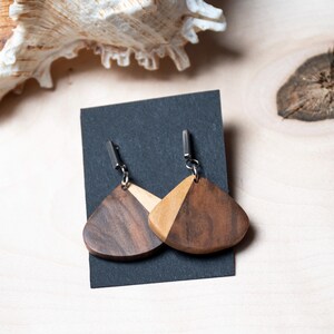 May include: A pair of wooden earrings with a light and dark wood design. The earrings are shaped like a teardrop and have a silver hook.